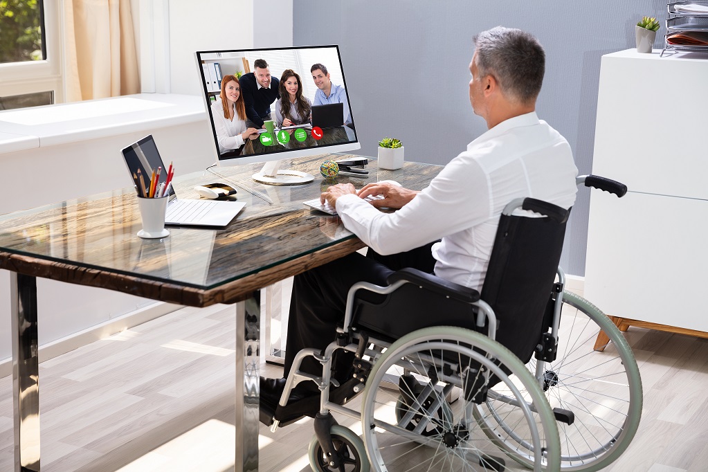 Foto de um homem grisalho, sentado em uma cadeira de rodas, com traje executivo. Acomodado em uma mesa de trabalho, ele tem as mãos sobre o teclado e olha para o monitor à sua frente, que exibe quatro pessoas em uma reunião virtual.