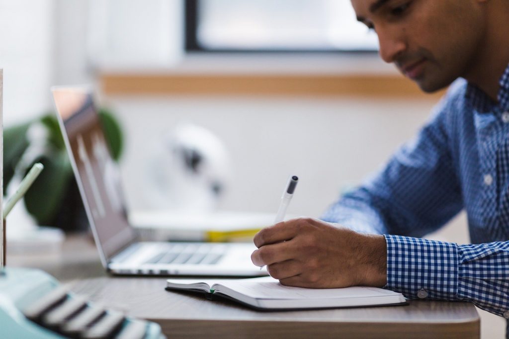 Foto de um homem sentado em uma mesa com laptop e caderno à sua frente. Ele segura uma caneta e olha para o caderno.