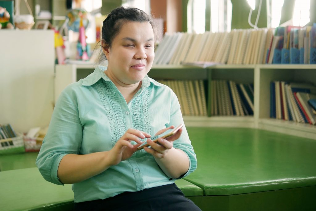 Foto em close de mulher cega em uma sala com prateleiras e vários livros. Ela segura um celular com a mão esquerda e toca a tela com os dedos da mão direita. Ela tem cabelos castanhos escuros, traços orientais e veste camisa verde clara. Ela sorri.