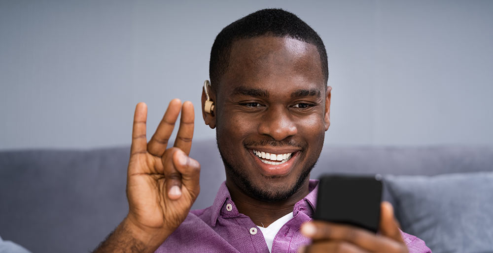 Foto de um homem negro com um aparelho auditivo, ele sorri olhando para o celular enquanto faz um sinal em Libras