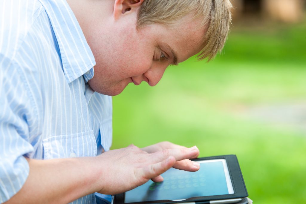 Foto de perfil de um jovem branco com cabelos loiros em ambiente externo. Ele olha atentamente para um tablet e toca a tela com seus dedos.