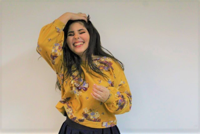 Foto da Jéssica Camarço em primeiro plano. Ela é uma jovem mulher de pele branca, cabelos castanhos escuros, lisos e compridos. Ela sorri com os lábios bem abertos, está com os olhos fechados e sua mão direita, no topo da cabeça, segura seus cabelos para trás. Ela veste veste camisa amarela com estampa floral. Está em um ambiente fechado com parede branca.