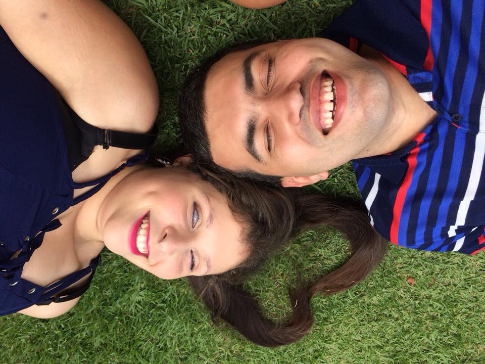 Foto de Leonardo Gleison e Camila Domingues deitados na grama em posições opostas e com suas cabeças lado a lado. Os cabelos da Camila estão ajeitados em formato de coração sobre a grama. Os dois sorriem de olhos fechados.