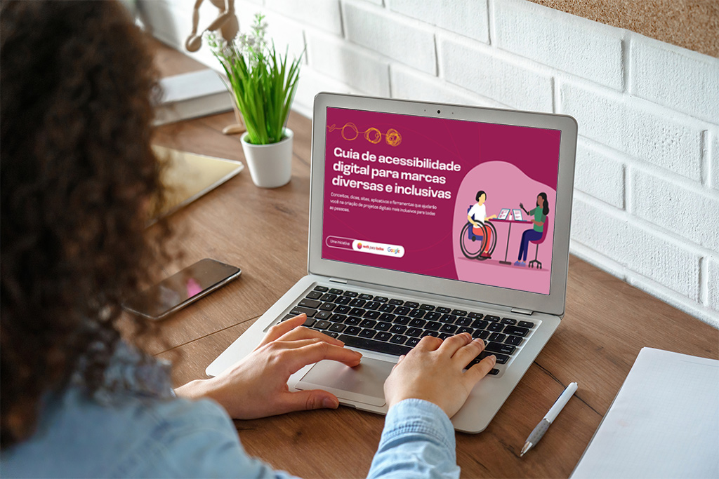 Foto de uma mulher de cabelos cacheados de costas usando um notebook. Na tela do aparelho, está a capa do Guia de acessibilidade digital para marcas diversas e inclusivas com a ilustração de duas mulheres, uma delas tem uma prótese em um dos braços.