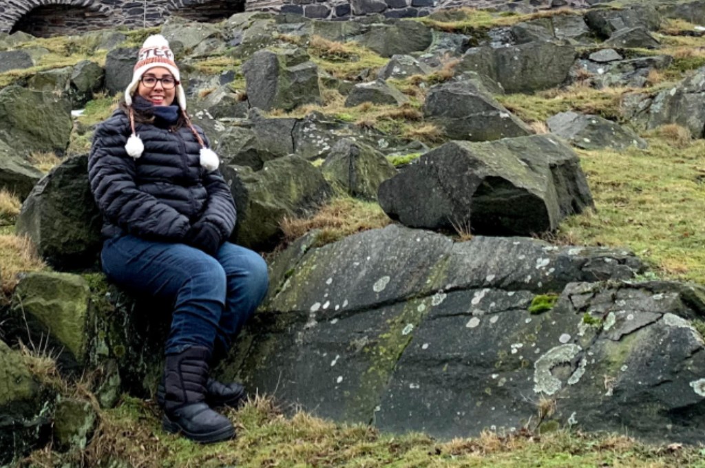 Foto de Marília Suzart sorrindo, sentada à frente de uma parede de pedras. Ela é branca, usa óculos de grau arredondado, veste jaqueta de frio, gorro, luvas e botas pretas.