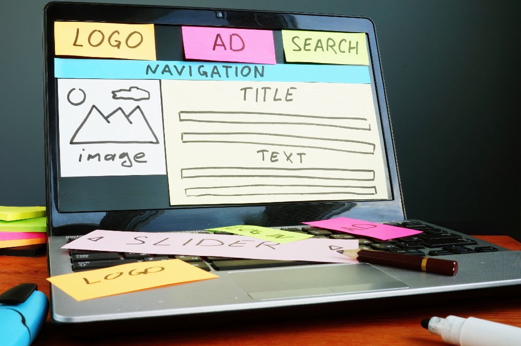 Foto de um notebook sobre uma mesa. Na tela do aparelho, há diversas notas autodesivas que repesentam as áreas de um site com os textos em inglês: "Logo, AD, Search, Navigation, Image, Title e Text".