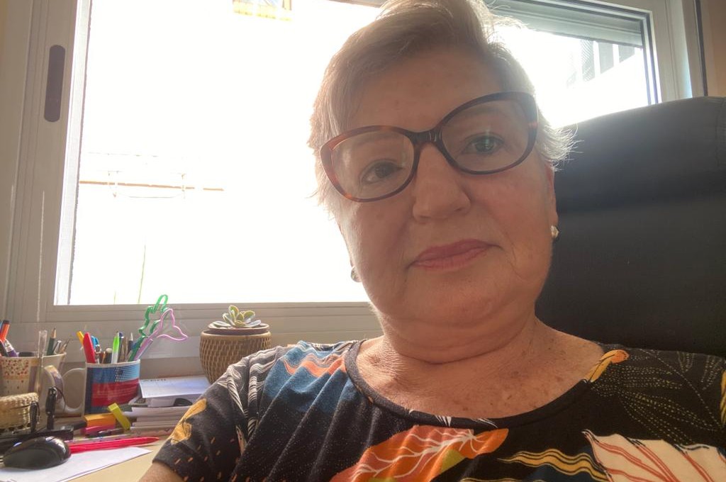 Foto estilo selfie de uma mulher branca, com cabelos loiros curtos, jogados para o lado direito. Ela usa óculos de grau com armação marrom, blusa preta florida e um par de brincos pequenos . Atrás dela, há itens de papelaria sobre uma mesa e uma grande janela.