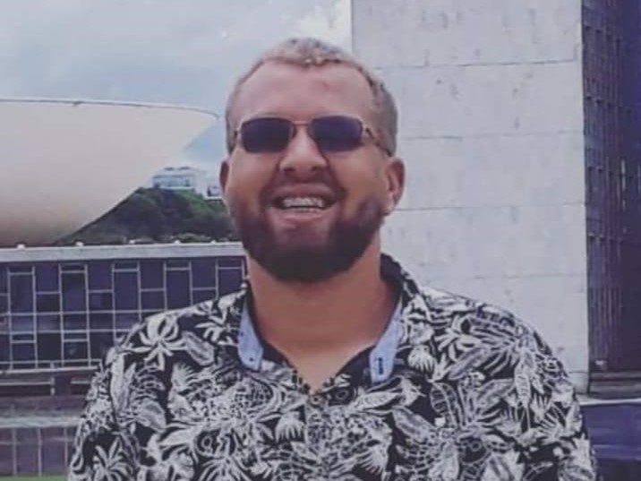 Foto em close do Carlos Barbosa. Ele é homem branco, cabelos curtos e loiros, barba e bigode castanhos claros. Usa óculos de sol com armação quadrada pequena e veste camisa preta estampada com com folhas brancas. Atrás dele há parte da fachada do Congresso Nacional, em Brasília.