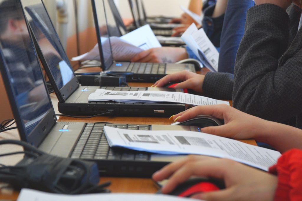 Foto de várias pessoas sentadas em frente a notebooks enfileirados sobre uma mesa de madeira. Na imagem é possível ver apenas parte de seus braços e mãos que tocam mouse, teclado e papeis.