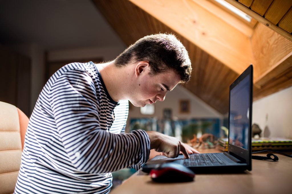 Foto de perfil de um jovem com síndrome de Down teclando em um notebook que está na mesa à sua frente. Ele é branco com cabelos castanhos curtos, veste camiseta longa branca listrada de preto e está sentado em uma cadeira.