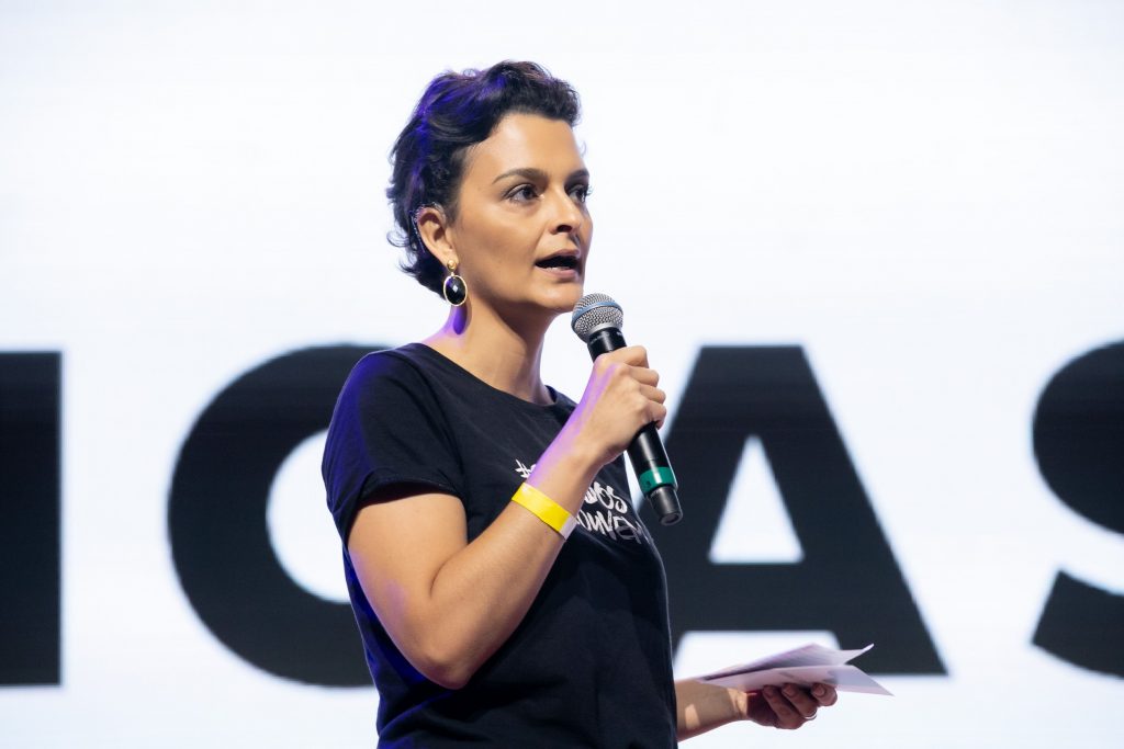 Foto de perfil da Paula Pfeifer. Ela é mulher branca de cabelos castanhos escuros bem curtinhos e olhos também castanhos. Ela segura um microfone perto de sua boca semiaberta e na outra mão uns papéis. Veste camiseta preta e atrás dela há umas letras pretas grandes estampadas numa parede branca.