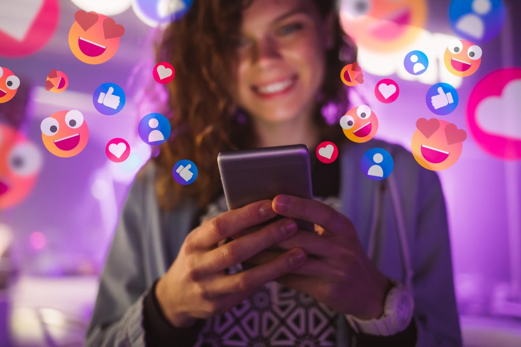 Arte com foto de mulher jovem que sorri enquanto olha para a tela do celular que segura em suas mãos. Há vários emojis relacionados às redes sociais sobrepostos na imagem.
