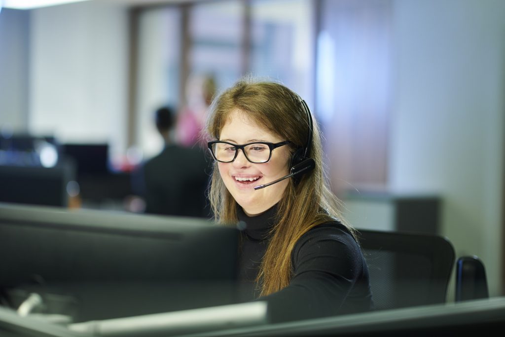 Foto de mulher jovem com Síndrome de Down sentada em frente a um computador. Ela usa óculos, veste camiseta preta, usa fone de ouvido com microfone e sorri.