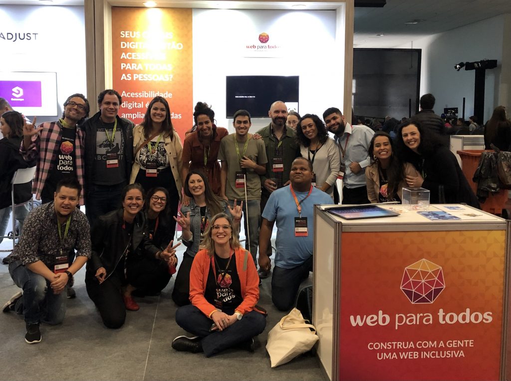 Foto tirada no estande do WPT no Digitalks. Há 17 pessoas bem juntinhas que sorriem. Elas estão atrás do pequeno balcão que apoia um notebook, brindes e cartilhas de acessibilidade. Nele há o logo do Movimento e o slogan "construa com a gente uma web inclusiva".