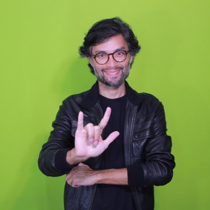 Foto de Alexandre Ohkawa, um homem de pele branca, cabelos castanhos, barba e bigode grisalhos e óculos de grau, em pé na frente de um fundo verde. Ele faz o sinal de eu te amo em Libras com a mão direita.