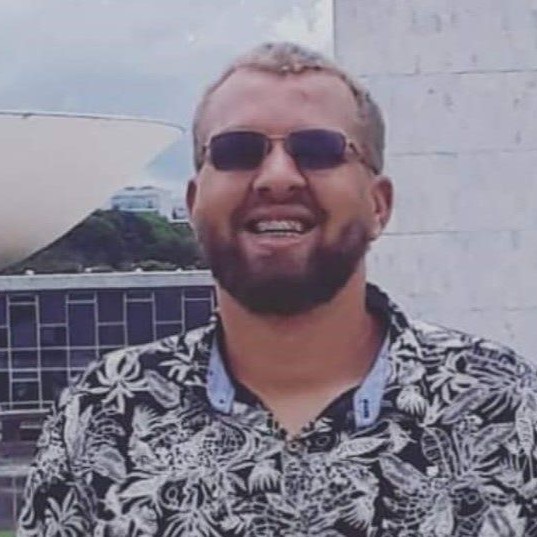 Foto em close do Carlos Barbosa. Ele é homem branco, cabelos curtos e loiros, barba e bigode castanhos claros. Usa óculos de sol com armação quadrada pequena e veste camisa preta estampada com com folhas brancas. Atrás dele há parte da fachada do Congresso Nacional, em Brasília.
