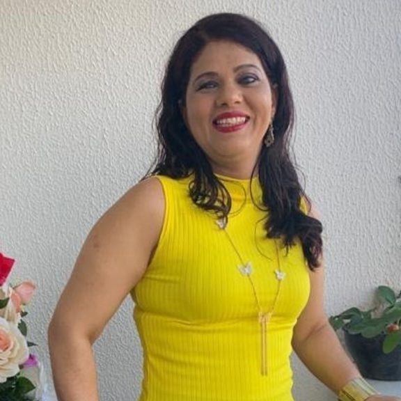 Doriane Vasconcelos em uma varanda. Ela é mulher branca e tem cabelos pretos e ondulados. Veste blusa amarela e usa batom vermelho.