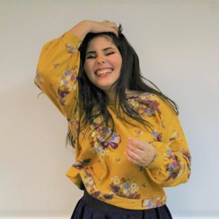 Foto da Jéssica Camarço em primeiro plano. Ela é uma jovem mulher de pele branca, cabelos castanhos escuros, lisos e compridos. Ela sorri com os lábios bem abertos, está com os olhos fechados e sua mão direita, no topo da cabeça, segura seus cabelos para trás. Ela veste veste camisa amarela com estampa floral. Está em um ambiente fechado com parede branca.
