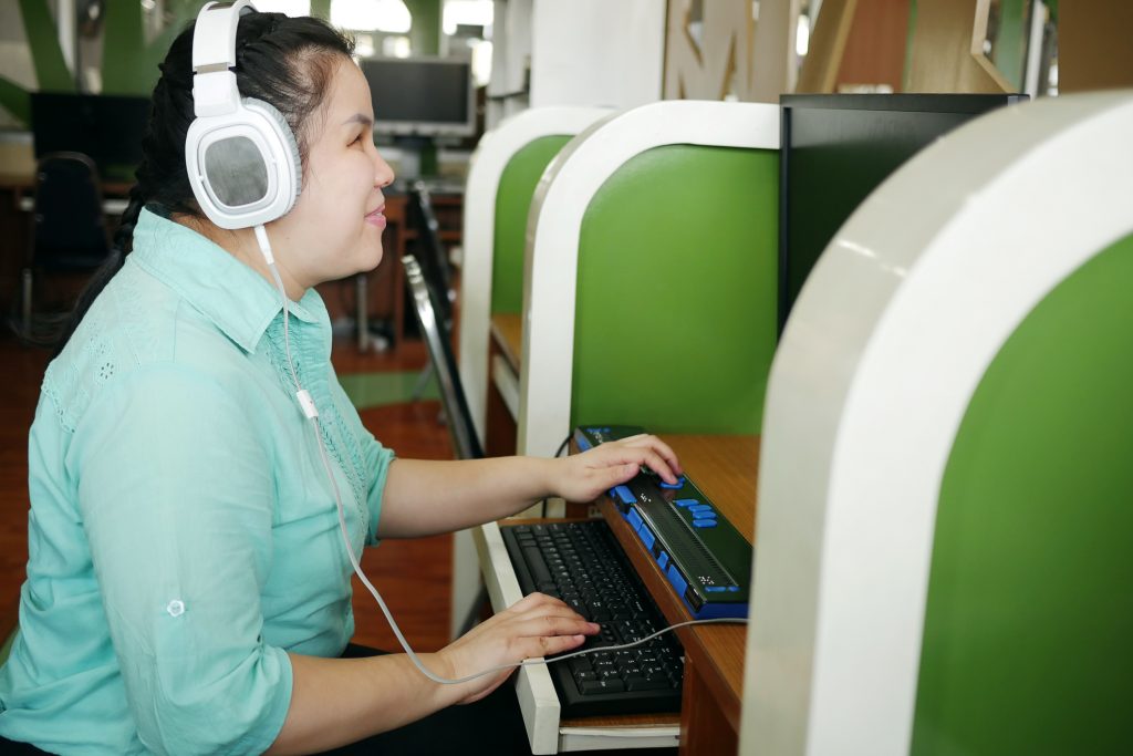 Mulher com características asiáticas apoia uma de suas mãos em um teclado comum de computador e a outra em um dispositivo Braille. Ela usa fones grandes nos ouvidos e sorri.