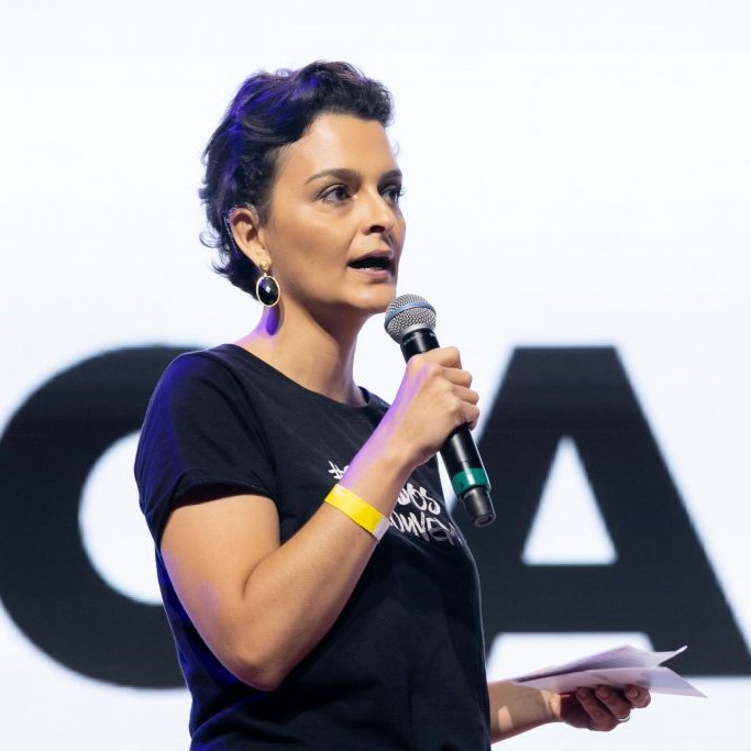 alt="Foto de perfil da Paula Pfeifer. Ela é mulher branca de cabelos castanhos escuros bem curtinhos e olhos também castanhos. Ela segura um microfone perto de sua boca semiaberta e na outra mão uns papéis. Veste camiseta preta e atrás dela há umas letras pretas grandes estampadas numa parede branca."