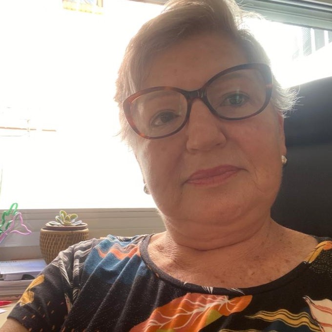 Foto estilo selfie de uma mulher branca, com cabelos loiros curtos, jogados para o lado direito. Ela usa óculos de grau com armação marrom, blusa preta florida e um par de brincos pequenos . Atrás dela, há itens de papelaria sobre uma mesa e uma grande janela.