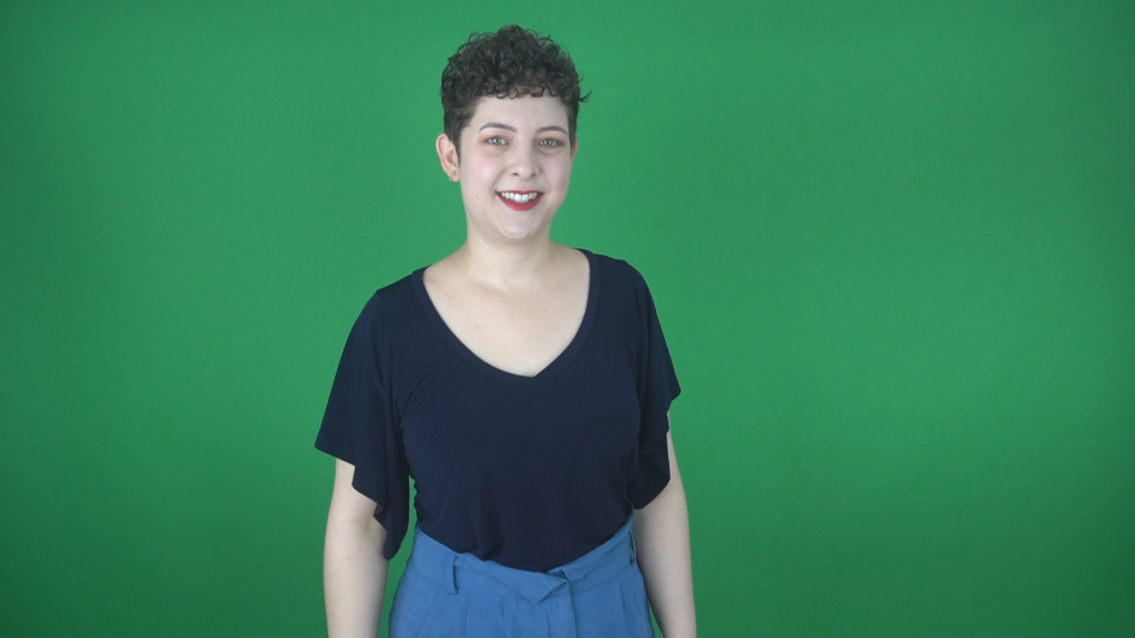 Foto em primeiro plano de Paloma Bueno em frente a uma parede verde. Ela é mulher branca, com olhos verdes claros e cabelos castanhos bem curtos e enrolados.