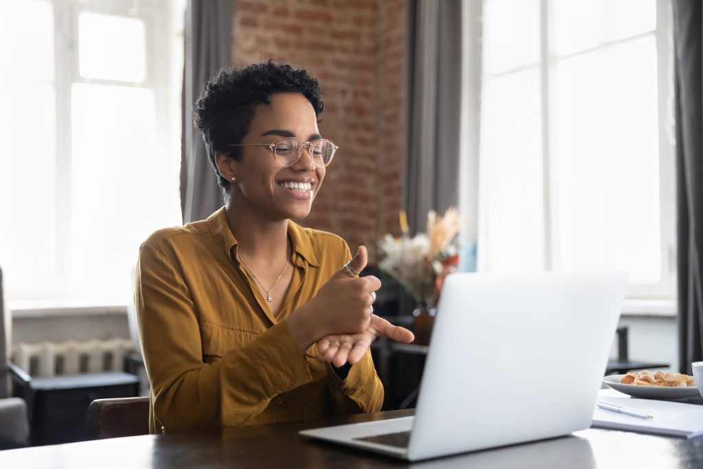 Foto de jovem negra sentada em frente a um notebook em um ambiente fechado com fundo desfocado. Ela olha e sorri para a tela do computador e faz um sinal de positivo com a mão esquerda fechada e o polegar para o alto, apoiada na palma da mão direita.