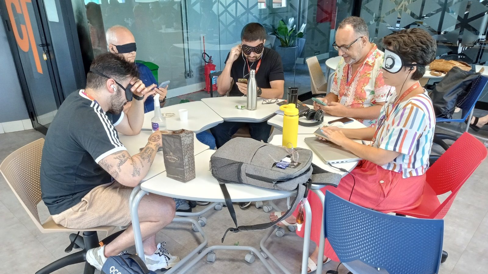 Foto panorâmica de sala de aula da CESAR School, com 5 estudantes em uma mesa redonda. Estão com os olhos vendados, utilizam fones de ouvido enquanto interagem com seus celulares em uma dinâmica de acessibilidade.