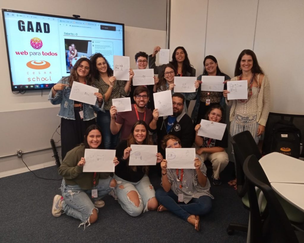 Foto de 13 jovens sorridentes no canto de uma sala de aula. Algumas pessoas estão em pé, outras agachadas e outras sentadas ou ajoelhadas no chão. Cada jovem segura uma folha de sulfite branca com desenhos feitos a lápis. Atrás do grupo há um telão onde há em destaque a palavra GAAD e os logos do Web para Todos e da Cesar School.