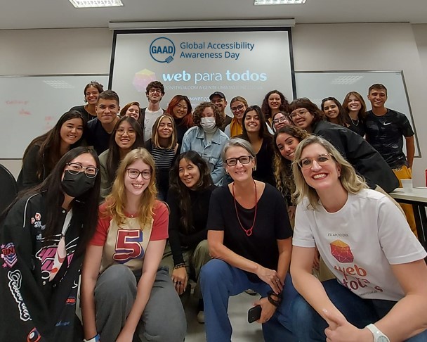 Foto de parte da turma de graduação em Design da ESPM São Paulo dentro da sala de aula. O grupo de 24 pessoas está bem juntinho, algumas estão em pé e outras abaixadas, todo mundo sorrindo. A professora Claudia Moura e Suzeli Damaceno estão no meio da turma. Os logotipos do Web para Todos e do Gaad estão projetados no telão atrás de todo mundo.
