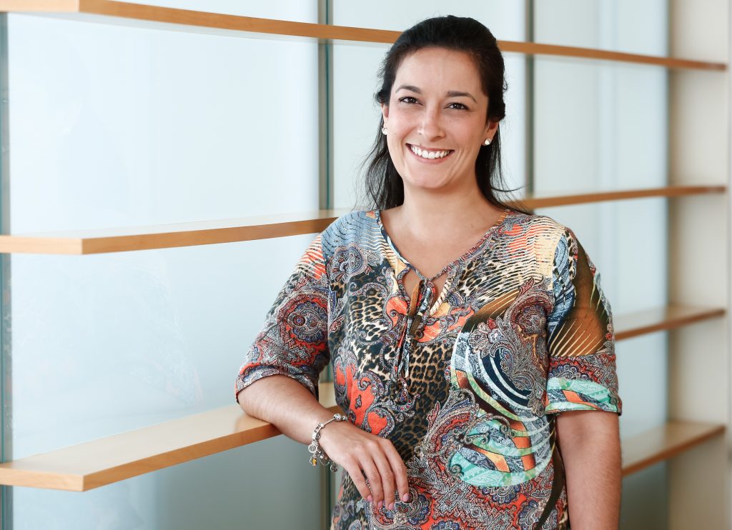 Foto de Luana Pablos em primeiro plano. Ela é branca, tem cabelos castanhos escuros, lisos e compridos, que estão presos para trás. Seu sorriso é largo e seus olhos também são castanhos. Veste uma camisa estampada e colorida e está apoiada em uma parede de vidro com prateleiras de madeira.