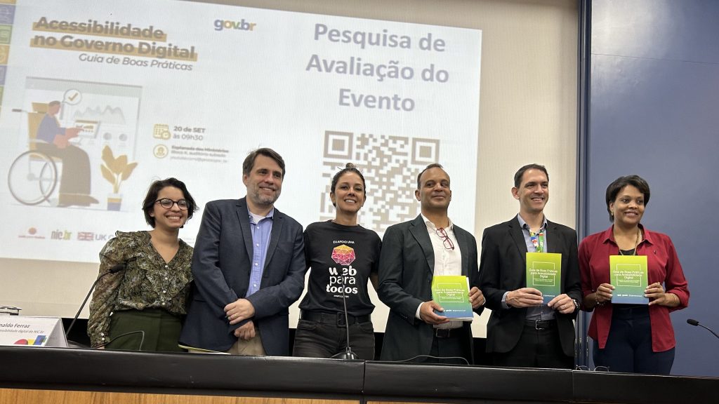 Foto de 6 pessoas enfileiradas, lado a lado, em frente a um painel com a projeção da arte do evento de lançamento do Guia de Boas Práticas em Acessibilidade Digital.