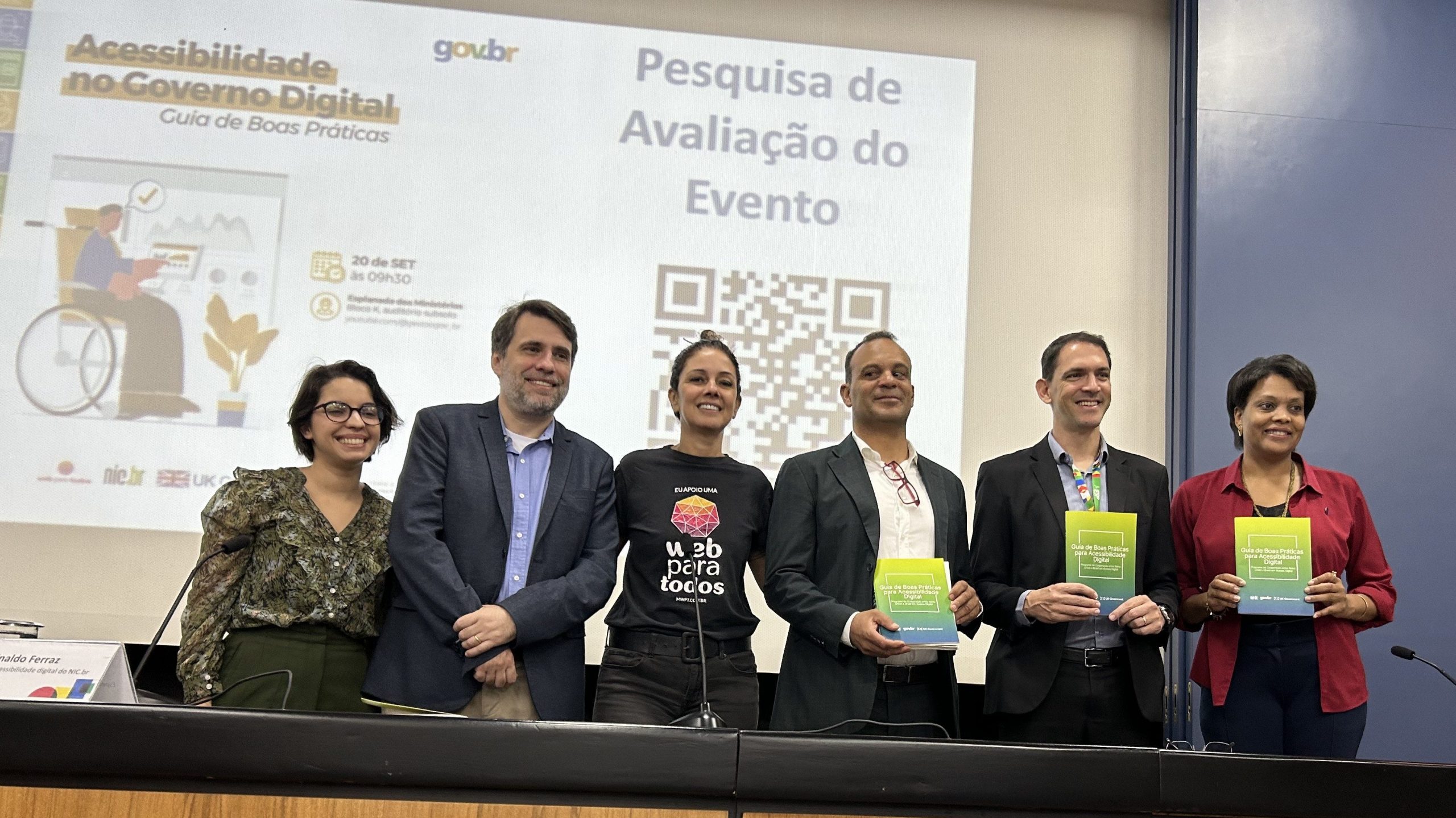 Foto de 6 pessoas enfileiradas, lado a lado, em frente a um painel com a projeção da arte do evento de lançamento do Guia de Boas Práticas em Acessibilidade Digital.