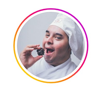 Foto de perfil de Gabriel Bernardes. Tem pele branca, olhos amendoados característicos da síndrome de Down. Veste roupa e chapéu de chef de cozinha e leva um brigadeiro à boca.