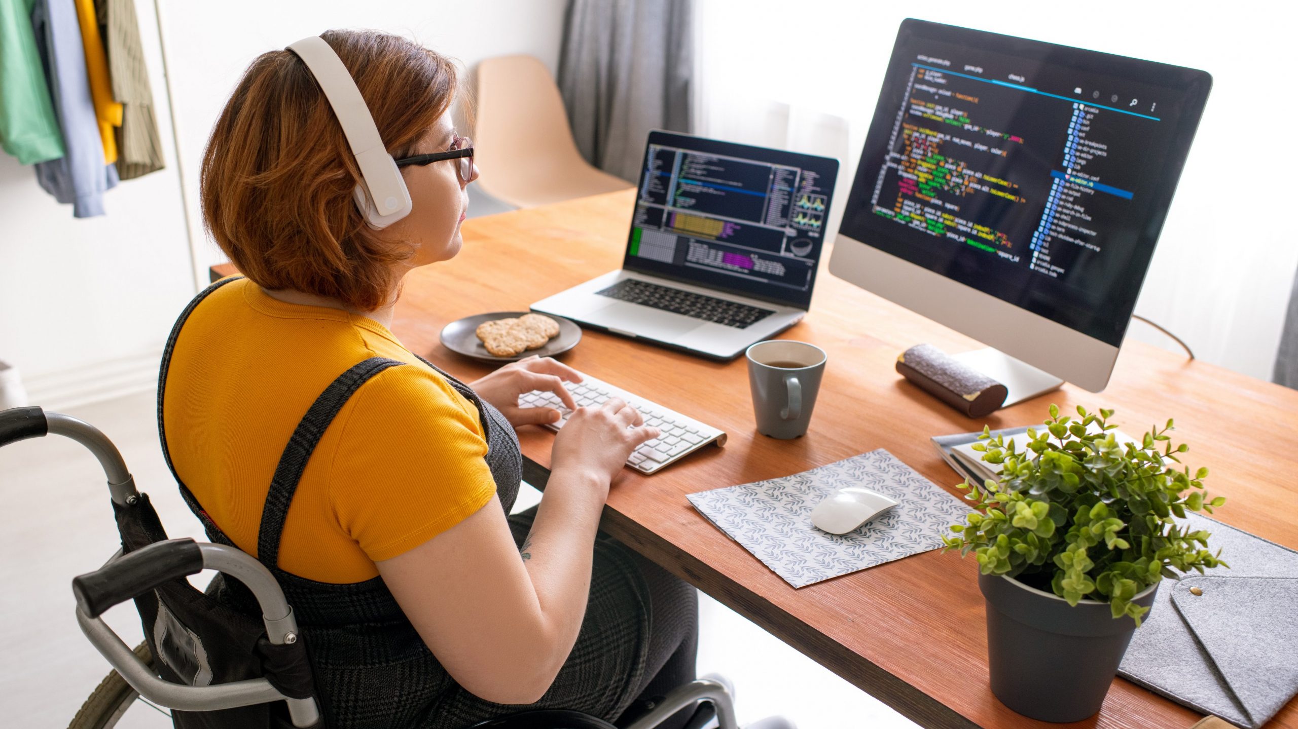 Foto de mulher cadeirante em frente a uma mesa com notebook, monitor grande, teclado móvel, mouse e outros objetos de escritório. Ela usa fone de ouvido e digita enquanto olha atentamente para a tela do monitor com códigos de programação projetados.