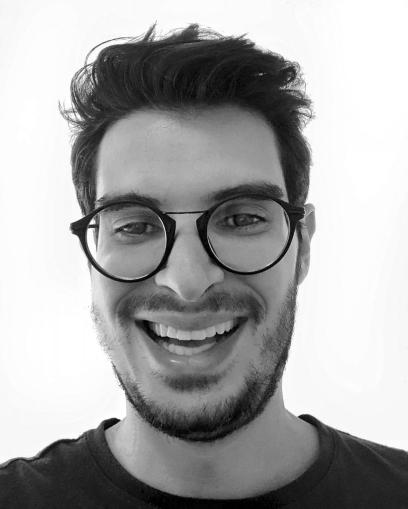 Foto em preto e branco de Diogo Cortiz sorrindo em close. Ele usa óculos e uma camiseta preta.