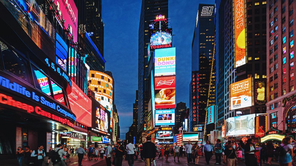 Foto da Times Square em Nova Yorque com muitos prédios, painéis iluminados e pessoas andando.