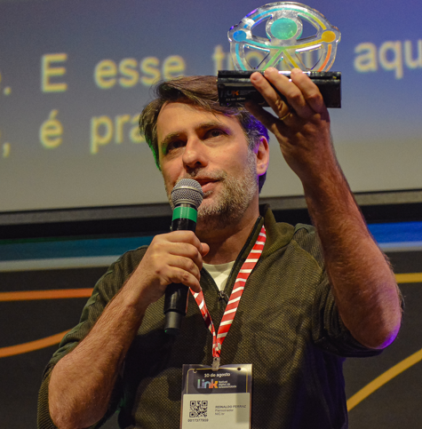 Foto de Reinaldo Ferraz em close. Ele segura um microfone e olha para frente. A outra mão segura um troféu.