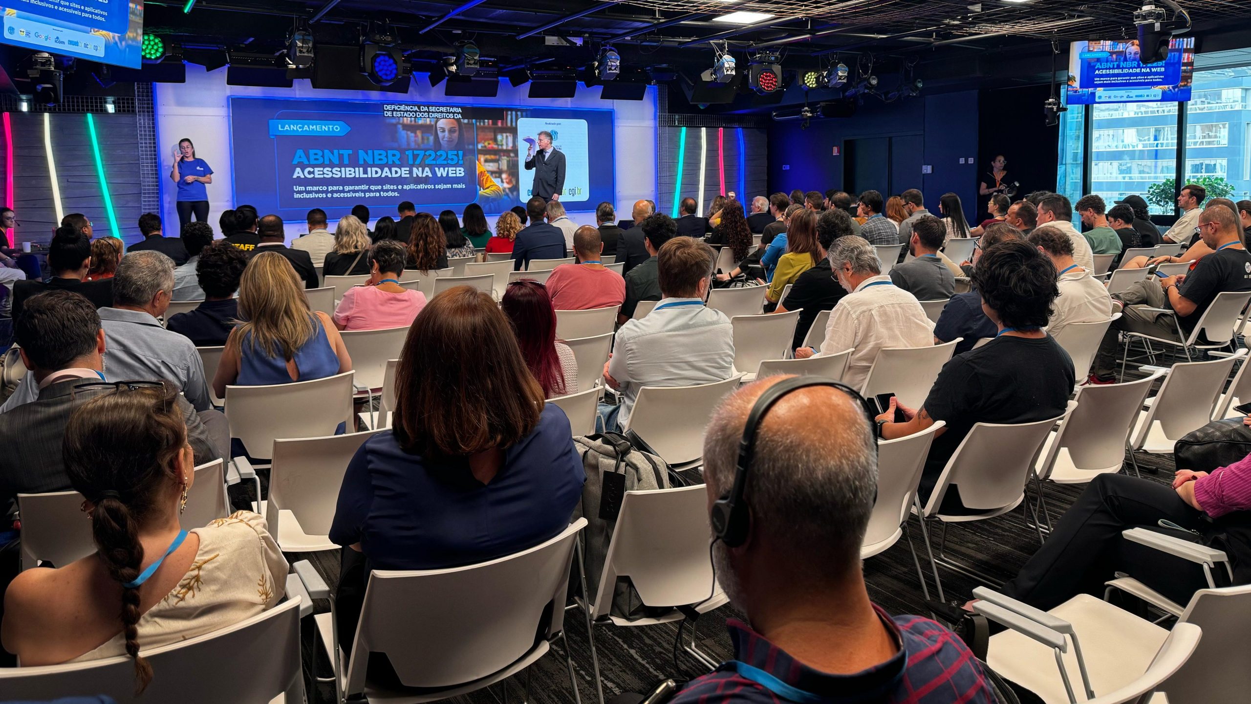 Foto do auditório tirada do fundo para o palco. Há dezenas de pessoas sentadas nas cadeiras olhando para o apresentador no palco, onde há um grande telão com o texto em destaque: "lançamento da ABNT NBR 17225. Acessibilidade na web". Há uma intérprete de Libras e legendas em tempo real projetadas no telão.