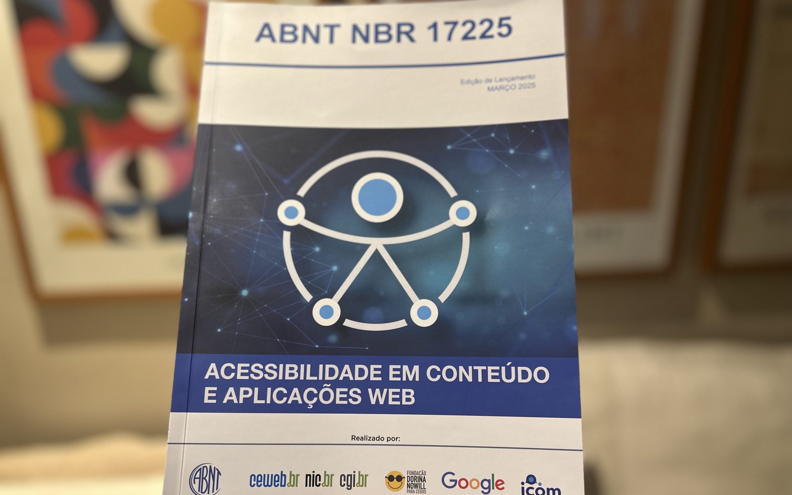 Foto da capa da edição especial de lançamento da NBR 17225. No centro dela há o símbolo universal da acessibilidade e o texto "ABNT NBR 17225. Acessibilidade em conteúdo e aplicações web. Realizado por: ABNT, Ceweb.br, Nic.br, CGI.br, Fundação Dorina Nowil, Google e Icom." O fundo da imagem está desfocado.