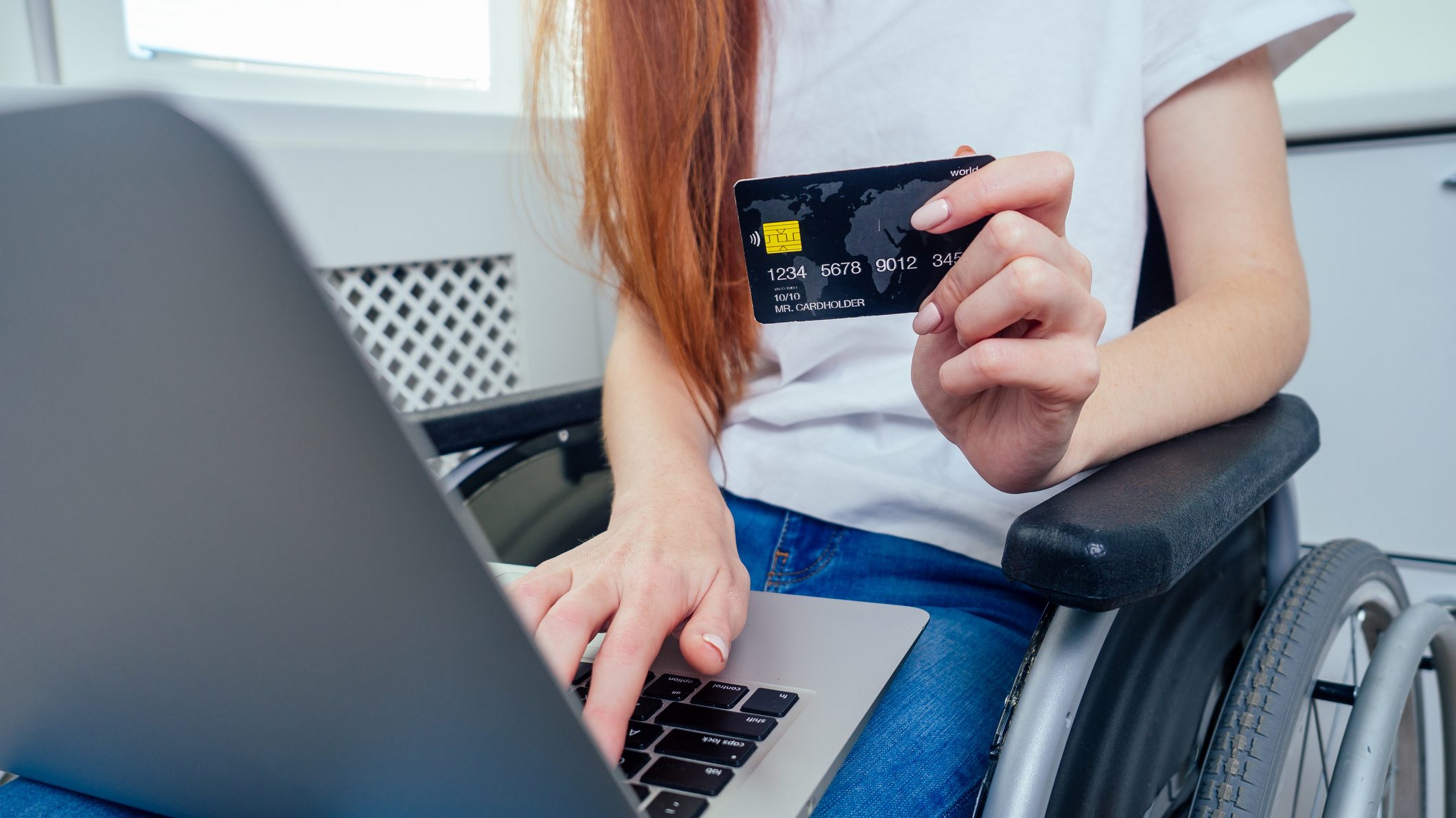 Foto de parte do corpo de uma jovem numa cadeira de rodas com um laptop sobre as pernas. Uma de suas mãos toca o teclado e a outra segura um cartão de crédito.