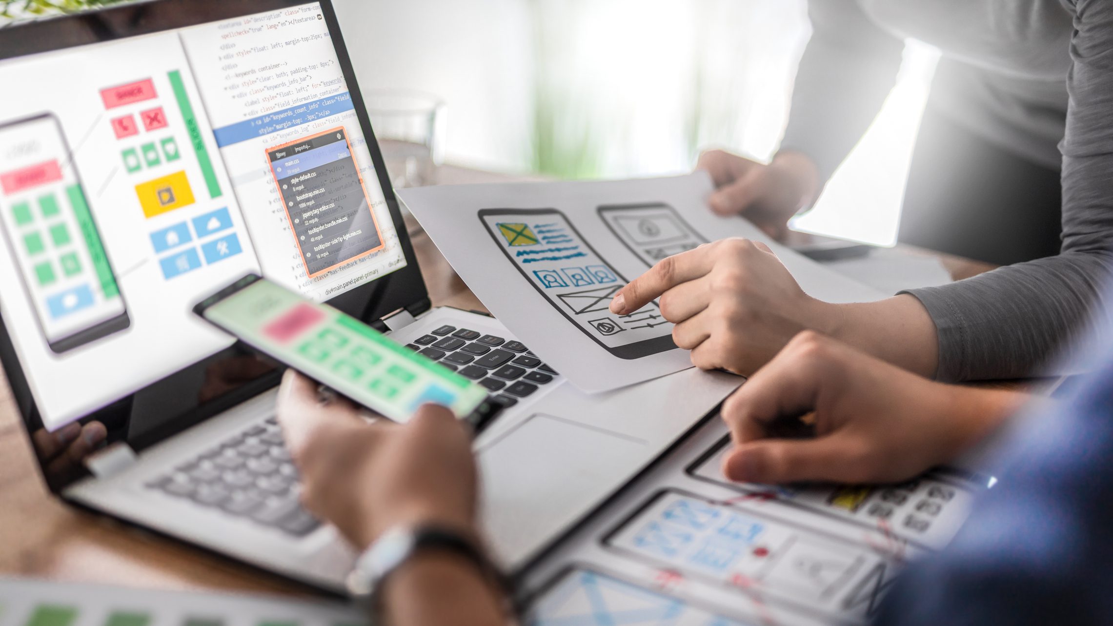 Foto de um ambiente criativo e colaborativo, com duas pessoas ao redor de um notebook. Elas ajustam elementos gráficos e textuais e revisam detalhes do projeto. Há símbolos ao redor que remetem ao conceito de webdesign.