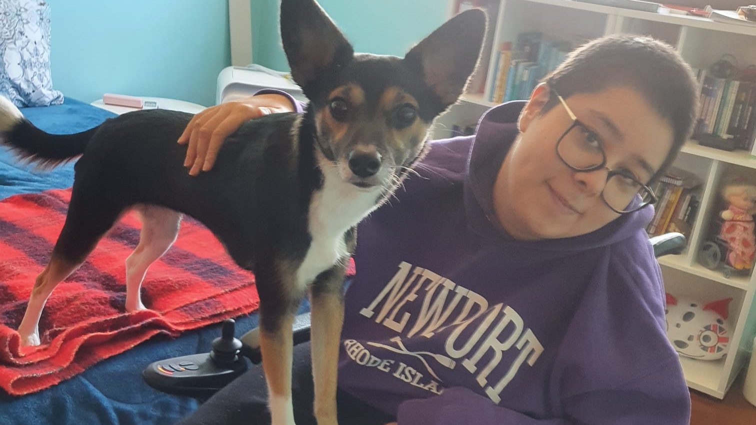 Foto de Paty Allerberger e seu cachorro em primeiro plano em um quarto. Ela é mulher jovem, branca, de cabelo curto, liso e castanho, olhos castanhos levemente e uma covinha na bochecha direita. Está sentada em sua cadeira de rodas.